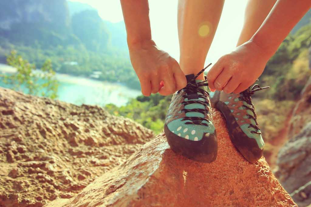 Zbliżenie na niebieskie buty wspinaczkowe. Kobieta próbuje je zawiązać stojąc na skale. W oddali widać rzekę i skały.